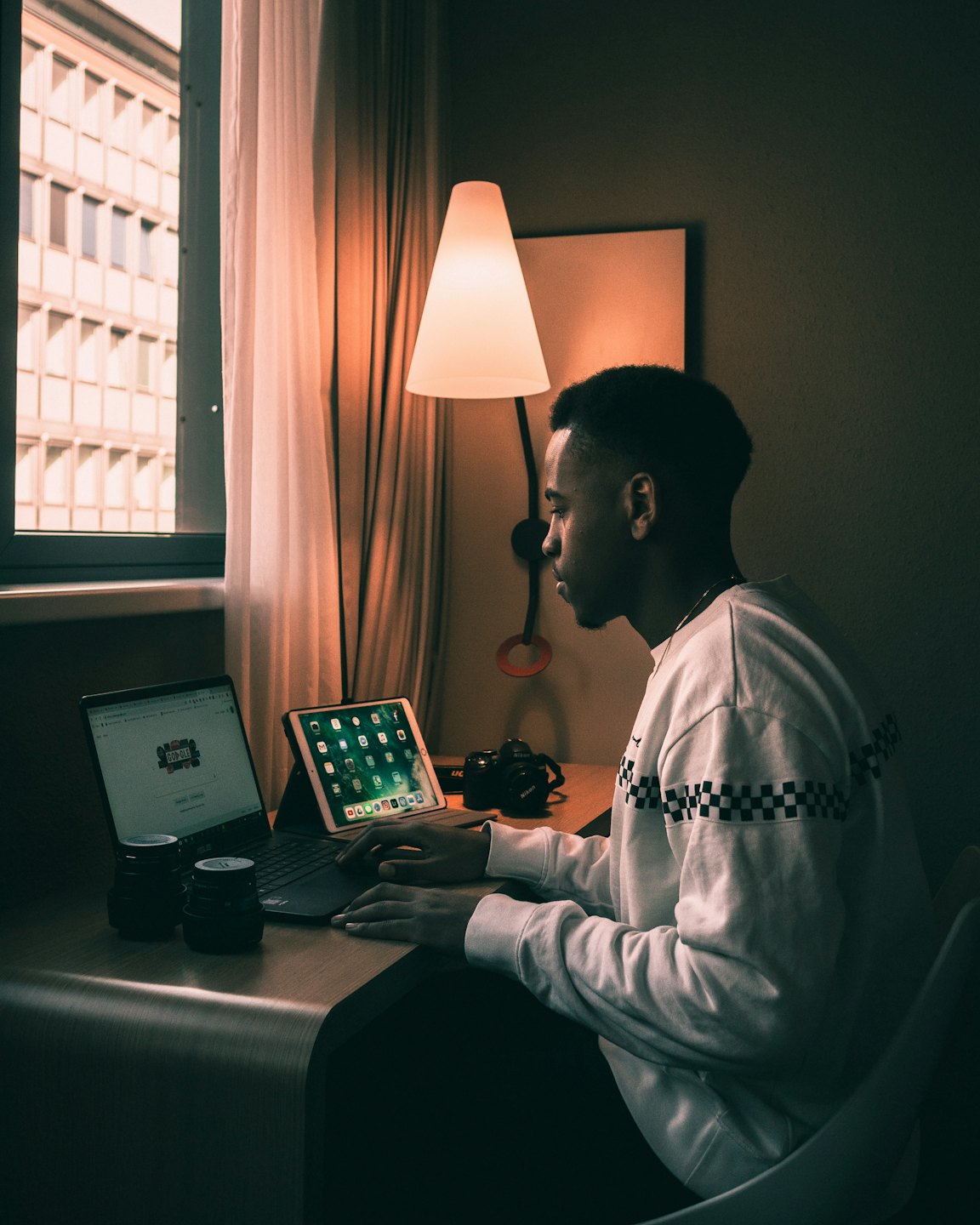 Man working on laptop and tablet at desk with camera equipment.