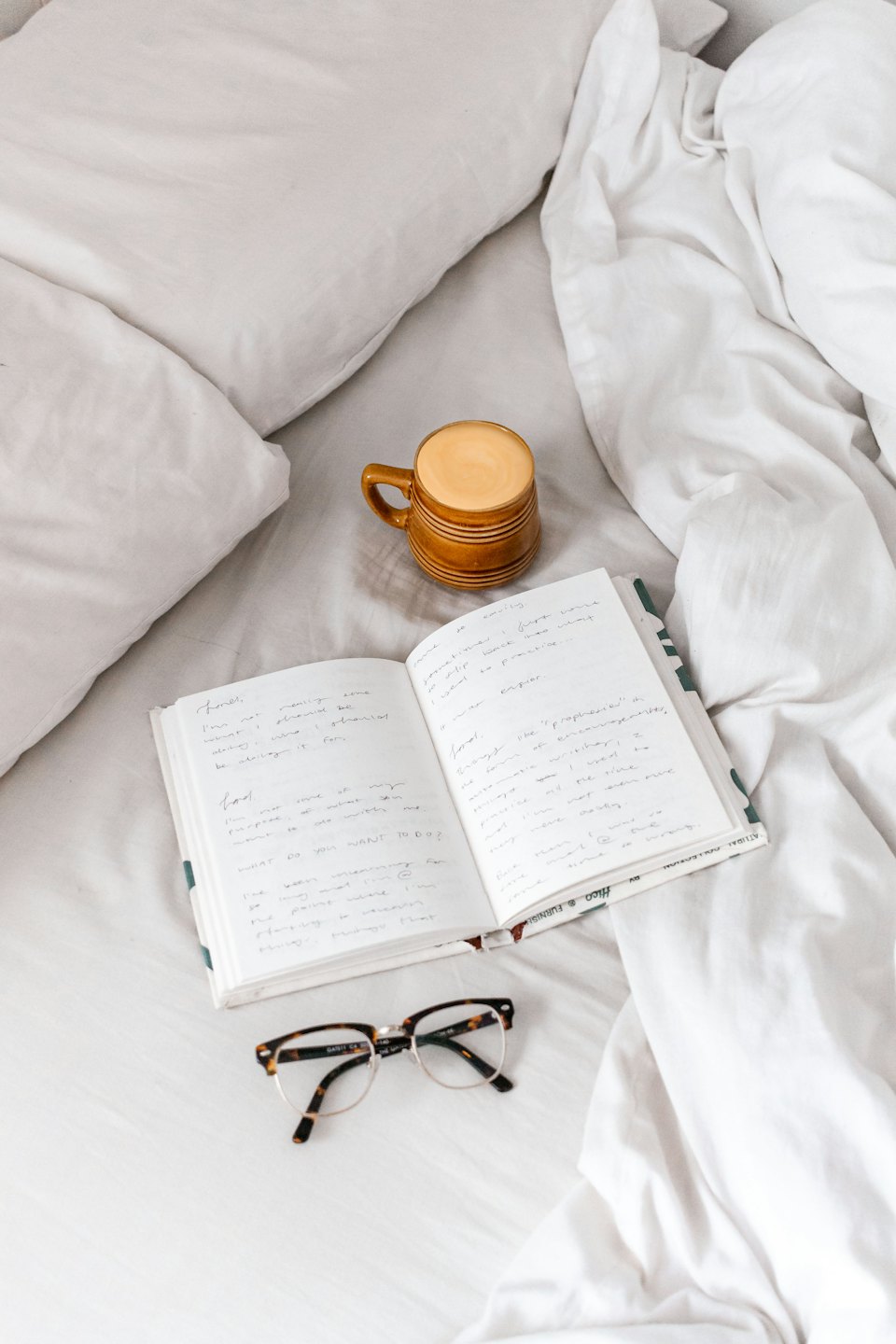 Journal, latte, and eyeglasses on rumpled white bed sheets.